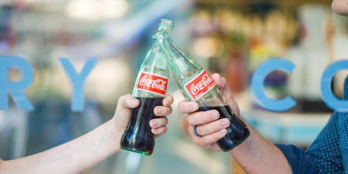 Two people, toasting with glass bottles of Coke