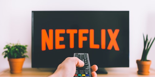 A hand holding a remote control in front of a television, turning on Netflix