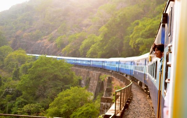 A train carrying a passenger (leaning out of the window) travels through green mountains