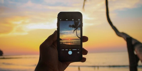 A hand holds a mobile phone taking a picture of a sunset on a beach