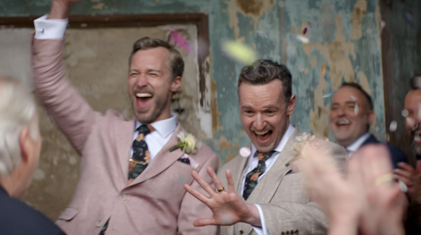 Two men in suits emerging from their wedding ceremony, one with his arm raised in celebration, the other looking at his wedding ring, as guests throw confetti