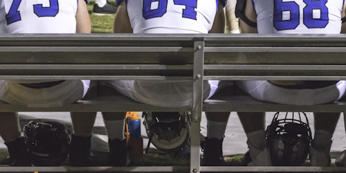 Three American football players sitting on sidelines bench with helmets between their feet